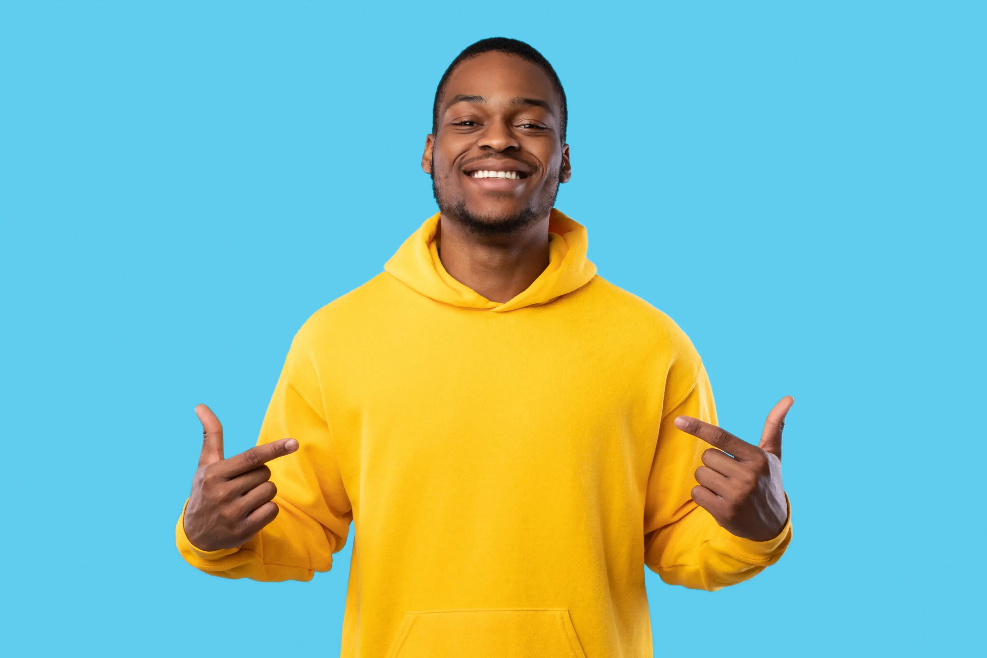 Cheerful Black Guy Pointing Fingers At Himself Over Blue Background Smiling person in yellow hoodie, blue background.